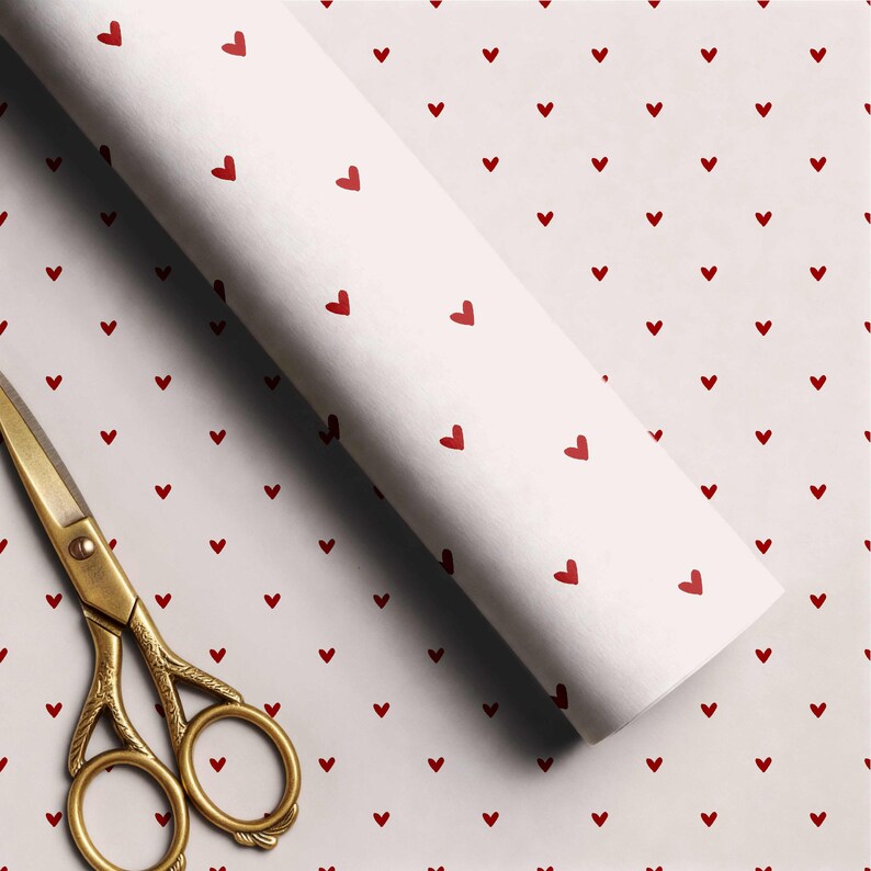May include: A roll of white wrapping paper featuring a repeating pattern of small red hearts, accompanied by a pair of gold-toned metal scissors with ornate handle detailing, positioned diagonally across the paper surface.