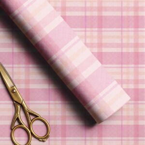 May include: A roll of wrapping paper featuring a pink, white, and light orange plaid pattern with a textured appearance, positioned diagonally across the frame next to a pair of ornate gold-colored metal scissors with intricate handle detailing.