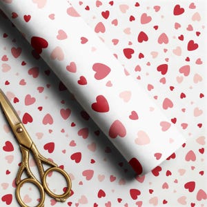 May include: A roll of white wrapping paper featuring a repeating pattern of scattered hearts in shades of red and light pink, positioned diagonally across the frame next to a pair of ornate gold-colored metal scissors with intricate handle details.