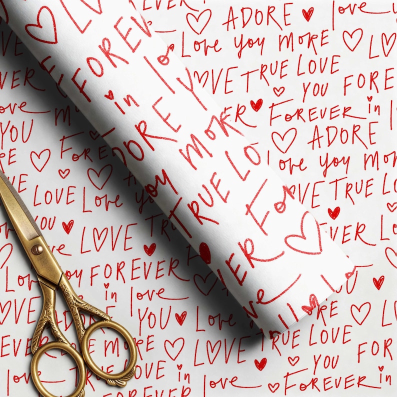 May include: A roll of white wrapping paper featuring a repeating red handwritten pattern of hearts and phrases including ADORE, Love you more, TRUE LOVE, and FOREVER. A pair of gold-toned metal scissors with ornate handles rests on the paper.