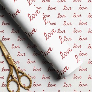 May include: A roll of white wrapping paper featuring a repeating pattern of the word love written in a dark red cursive script, accompanied by a pair of ornate gold-colored metal scissors with intricate detailing on the handles and blades.