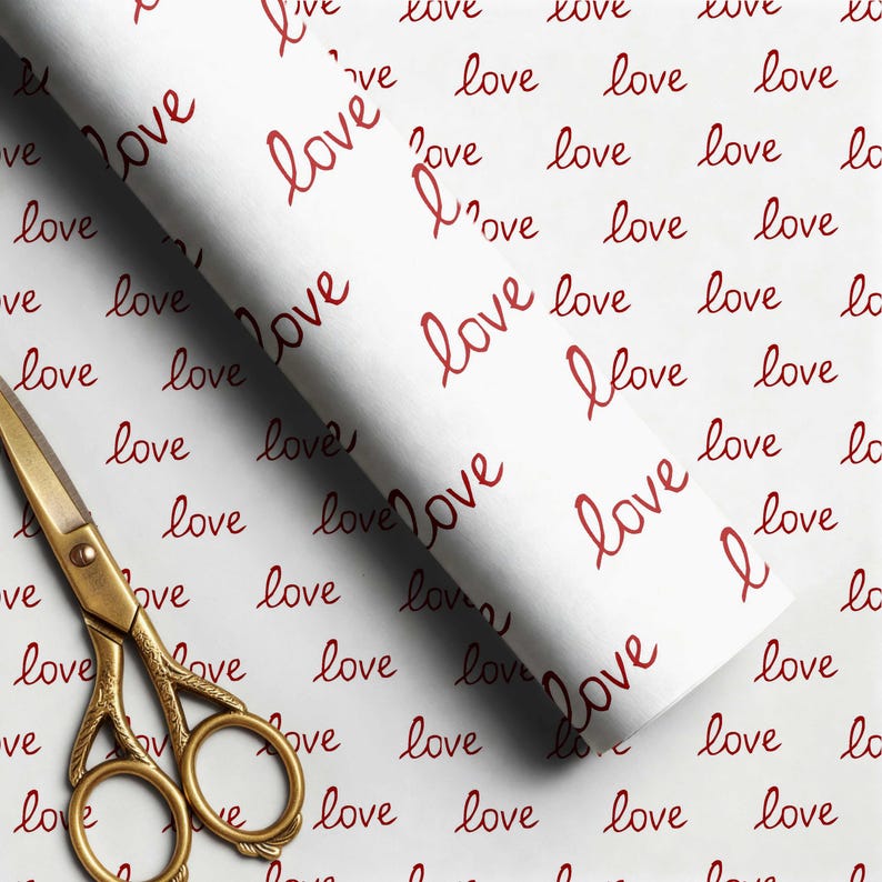May include: A roll of white wrapping paper featuring a repeating pattern of the word love written in a dark red cursive script, accompanied by a pair of ornate gold-colored metal scissors with intricate detailing on the handles and blades.