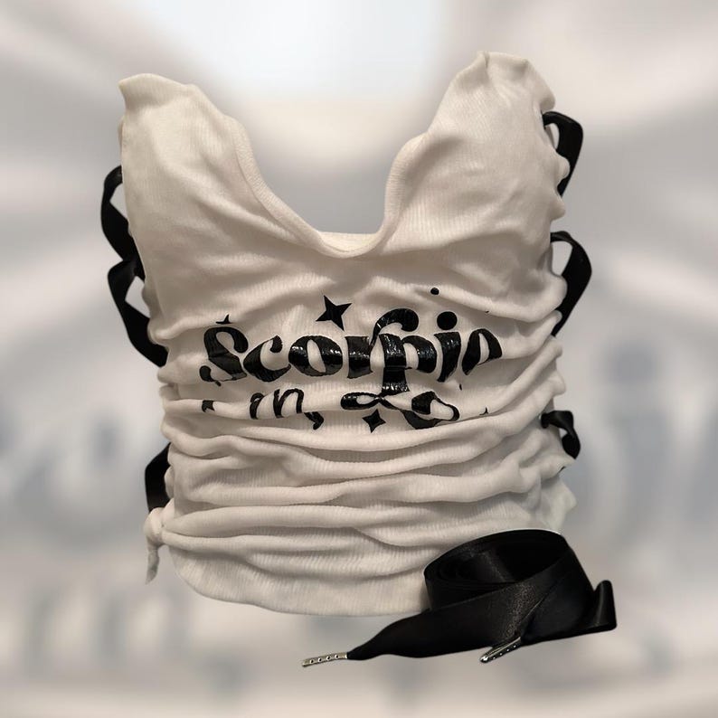 May include: White ruched top with black text that reads "Scorpio in". The top has black ribbon detailing and a black ribbon drawstring. The top is a sleeveless design.