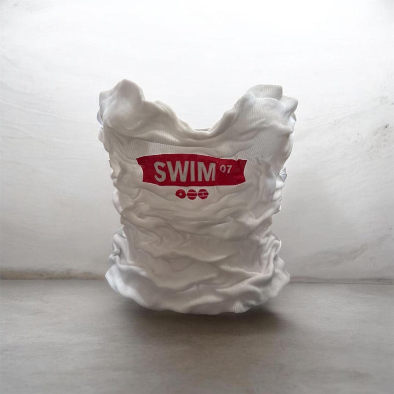 May include: A white, textured sculpture resembling a crumpled t-shirt. The red text on the shirt reads "SWIM '07" above three red circles. The sculpture is set against a light gray background.
