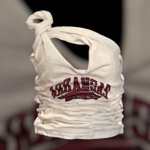 May include: White tank top with the word "ARKANSAS" in maroon lettering and a maroon Razorback logo. The top is gathered and knotted at the shoulder, with a scoop neckline.