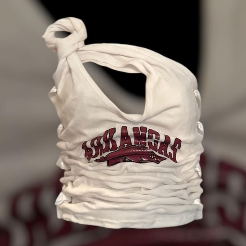 May include: White tank top with the word "ARKANSAS" in maroon lettering and a maroon Razorback logo. The top is gathered and knotted at the shoulder, with a scoop neckline.