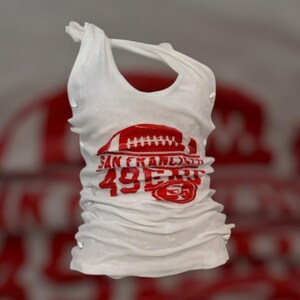 May include: White tank top with a red football graphic and the text "SAN FRANCISCO 49ERS". The top has a halter neck design and is made of a soft, lightweight fabric. The graphic includes the team logo.
