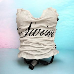 May include: White ruched top with the word "Summer" in black script. The top has a scoop neck and a black ribbon detail at the bottom. The background is a gradient of blue and pink.
