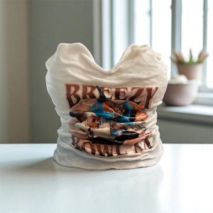 May include: A white ceramic sculpture in the shape of a t-shirt. The shirt has the word "BREEZY" in brown letters and a colorful image of people. The sculpture is on a white surface.