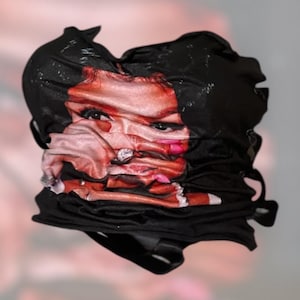May include: Black fabric top with a printed face design. The face has red lips, dark eyes, and a light skin tone. The fabric is gathered and ruched, creating a textured appearance. The top has a strapless design.