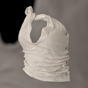 May include: A white, ruched halter top with a knotted strap. The top features a series of silver-colored eyelets along the side. The fabric has a ribbed texture, and the overall design is minimalist and modern.