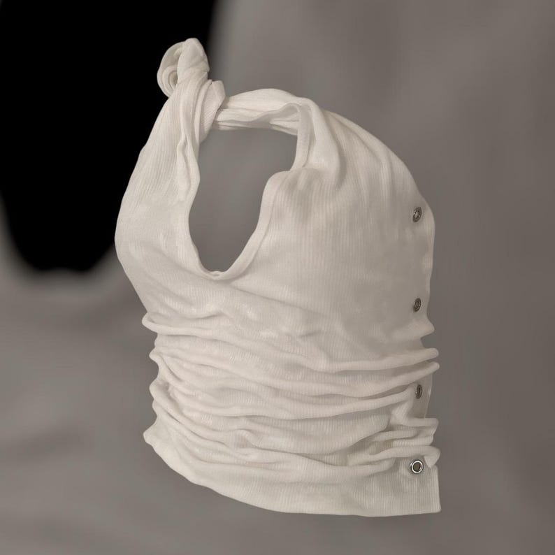 May include: A white, ruched halter top with a knotted strap. The top features a series of silver-colored eyelets along the side. The fabric has a ribbed texture, and the overall design is minimalist and modern.