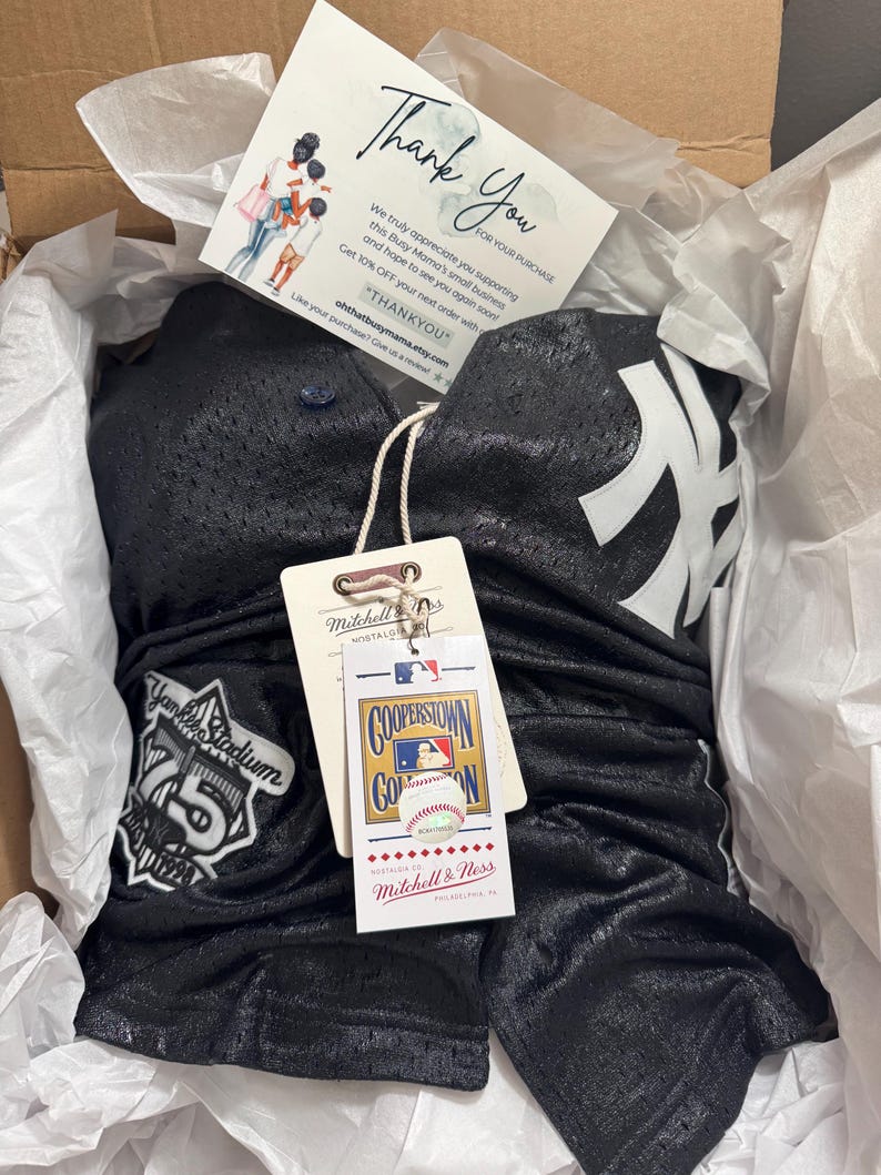 May include: A black mesh baseball jersey and shorts set with white accents, featuring the New York Yankees logo. The set is packaged in a box with a thank you card and a Mitchell & Ness tag.