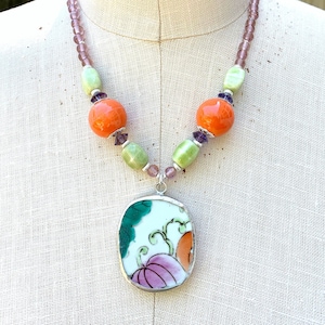 May include: A necklace with a large oval pendant featuring a floral design in shades of pink, orange, and green. The pendant is made from a piece of vintage china. The necklace is made with orange, green, and purple beads.