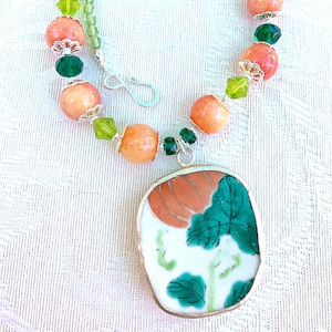 Puede incluir: Un collar de cuentas de color melocotón y verde con un colgante que presenta un diseño floral sobre un fondo blanco.