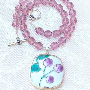 May include: A necklace with a silver pendant featuring a vintage porcelain design with purple and green floral motifs. The necklace is made with light pink faceted glass beads.