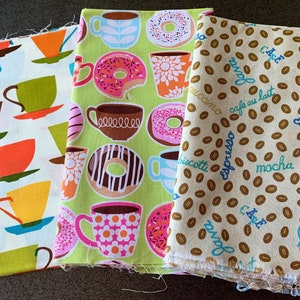 May include: Three fabric panels with different patterns. The first panel has a white background with colorful teacups and saucers. The second panel has a green background with donuts and teacups. The third panel has a beige background with brown coffee beans and the words "cafe", "espresso", "latte", "mocha", "biscotti", "cappuccino", and "cafe au lait" in blue.
