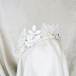 May include: White floral crown with delicate leaves and pearl accents. The crown is made of white metal and has a delicate, feminine design.