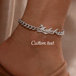 May include: A silver-toned curb chain anklet featuring a central nameplate with the custom text "Thaleek" in a cursive script. The nameplate is accented with a small crown icon above the first letter and a heart shape below the name.