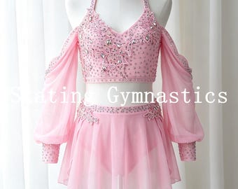 May include: A light pink two-piece skating and gymnastics outfit featuring a halter-style crop top and a matching skirt. The top is adorned with intricate floral lace appliques and scattered clear crystals. It includes long, sheer, billowy sleeves.