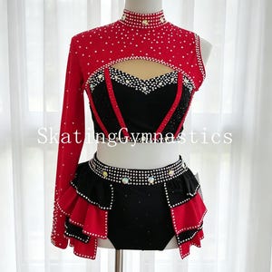 Puede incluir: Traje de patinaje o gimnasia rojo y negro con pedrería. La parte superior tiene cuello alto y una manga larga. La falda tiene un diseño con volantes. El texto "Skating Gymnastics" es visible.