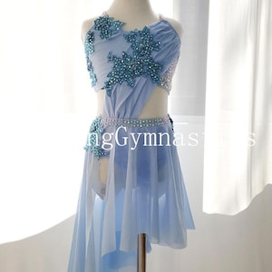 May include: A light blue dance costume with a cut-out design and asymmetrical skirt. The bodice is embellished with sparkling blue floral appliqués and a beaded belt. The costume is displayed on a mannequin.