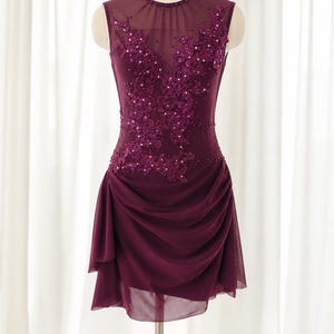 May include: A sleeveless, deep plum-colored dress featuring a sheer mesh neckline, intricate floral lace appliques with matching sequins, and a gathered, layered skirt that falls above the knee, displayed on a dress form mannequin.