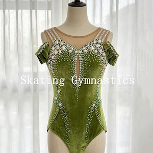 May include: An olive green velvet leotard featuring a nude mesh neckline, off-the-shoulder sleeves, and intricate crystal embellishments in various shapes and sizes. The garment displays the text Skating Gymnastics across the centre front.
