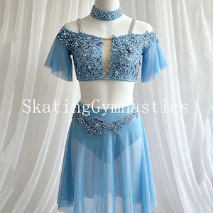 May include: A light blue two-piece performance outfit featuring a cropped top with off-the-shoulder sleeves, a choker, and a skirt. The set is adorned with intricate floral lace, silver sequins, and clear crystals. The text SkatingGymnastics is visible.