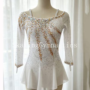 May include: A white long-sleeved leotard featuring a scoop neckline, thin shoulder straps, and a flared skirt. The bodice is embellished with a dense pattern of clear and amber-colored crystals arranged in diagonal lines. The text SkatingGymnastics is visible.