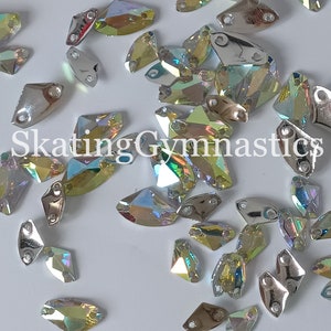 May include: A large assortment of iridescent, faceted, clear glass rhinestones with silver settings. The rhinestones are scattered on a white surface. The rhinestones are in various shapes and sizes. The text "Skating Gymnastics" is visible in the image.