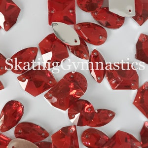 May include: Assortment of red crystal rhinestones in various shapes, including ovals, squares, and teardrops. The rhinestones are scattered on a white surface.