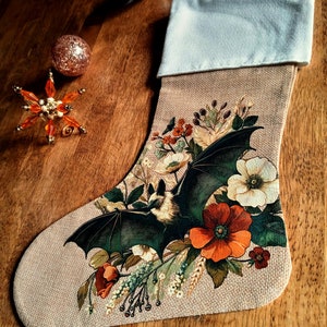 May include: A beige burlap Christmas stocking with a white cuff and a hanging loop. The stocking features a printed graphic of a bat with dark wings surrounded by a colorful arrangement of orange, red, and white flowers and green foliage.