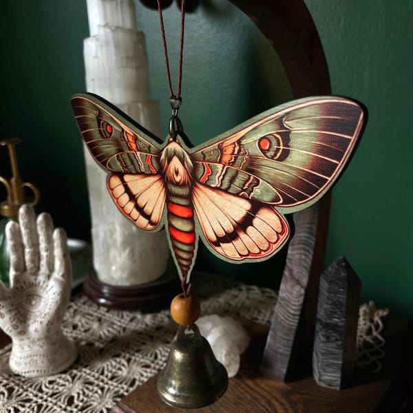 May include: A wooden moth ornament with green and orange wings, a red and orange body, and a small brass bell. The moth is suspended by a brown string. The ornament is a decorative item.