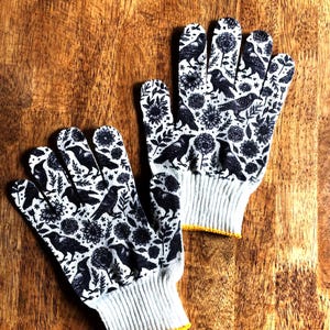Op de afbeelding: Een paar witte werkhandschoenen met een herhalende zwarte grafische print van kraaien en diverse bloemmotieven. De handschoenen hebben een geribbelde gebreide boord met een gele zoom, wat zorgt voor een goede pasvorm tijdens het werken.
