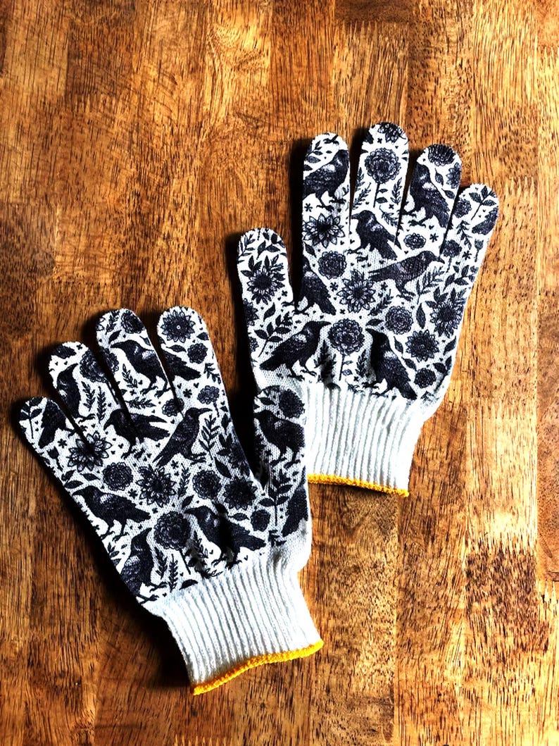 Op de afbeelding: Een paar witte werkhandschoenen met een herhalende zwarte grafische print van kraaien en diverse bloemmotieven. De handschoenen hebben een geribbelde gebreide boord met een gele zoom, wat zorgt voor een goede pasvorm tijdens het werken.
