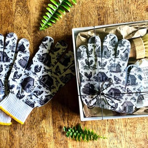 Op de afbeelding: Een paar witte handschoenen met een zwarte print van kippen en hokken, naast een geschenkdoos met een bijpassend paar handschoenen, een houten schrobborstel en een stuk Dirty Bird Oats & Honey zeep van 114g, alles verpakt in bruin papier.