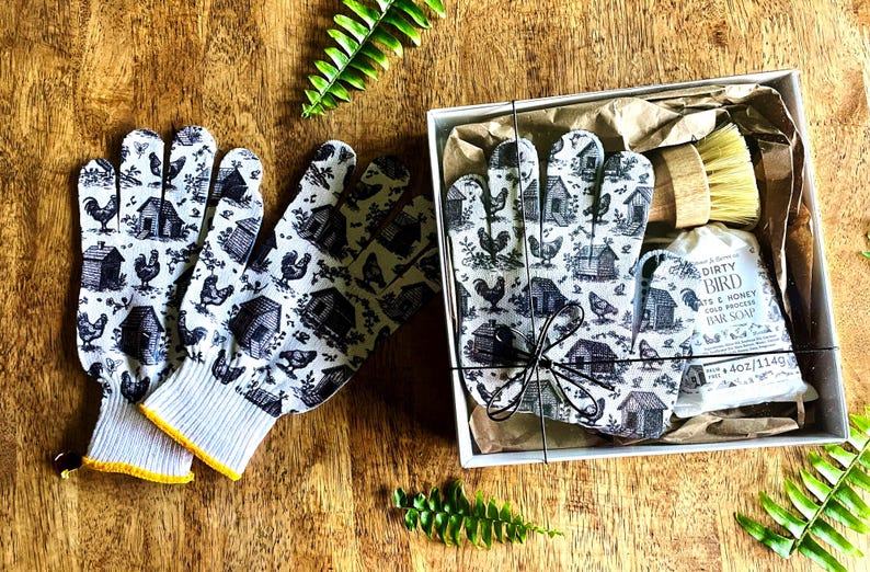 Op de afbeelding: Een paar witte handschoenen met een zwarte print van kippen en hokken, naast een geschenkdoos met een bijpassend paar handschoenen, een houten schrobborstel en een stuk Dirty Bird Oats & Honey zeep van 114g, alles verpakt in bruin papier.