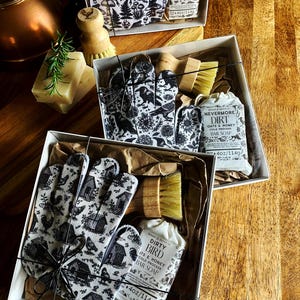 Op de afbeelding: Drie geschenkdozen met schoonmaaksets, elk met een handschoen met patroon, een houten schrobborstel en een stuk zeep van 114g. De zeepetiketten bevatten de teksten Toad-Ally Clean, Nevermore Dirt en Dirty Bird met illustraties.