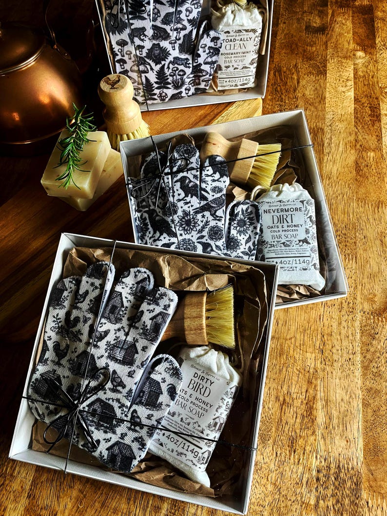 Op de afbeelding: Drie geschenkdozen met schoonmaaksets, elk met een handschoen met patroon, een houten schrobborstel en een stuk zeep van 114g. De zeepetiketten bevatten de teksten Toad-Ally Clean, Nevermore Dirt en Dirty Bird met illustraties.