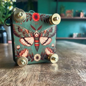 May include: A decorative box with a teal background featuring a moth illustration with red and white accents. The box has brass hardware and floral details. The box is sitting on a wooden surface.