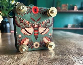 Op de afbeelding: Een decoratieve doos met een turquoise achtergrond met een motillustratie met rode en witte accenten. De doos heeft messing hardware en bloemendetails. De doos staat op een houten oppervlak.