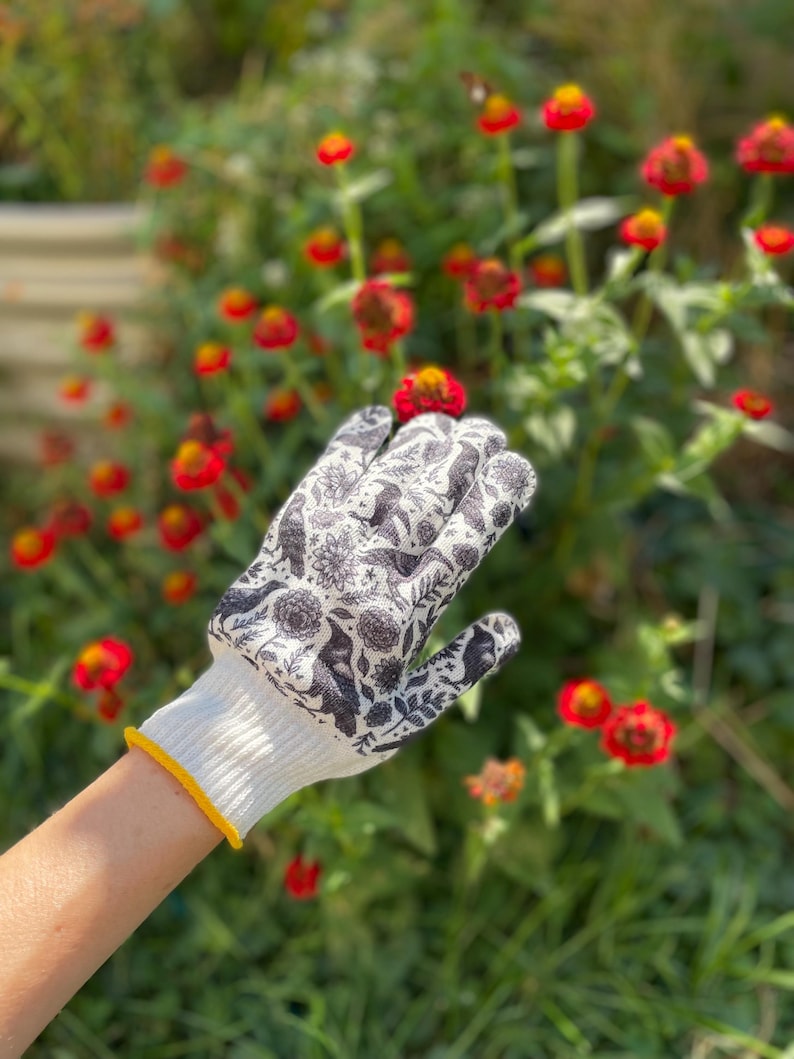 Op de afbeelding: Een witte tuinhandschoen met een zwarte botanische print en vogelmotieven. De handschoen heeft een geribbelde witte boord met een dunne gele rand, ontworpen voor handbescherming tijdens het werken met planten en bloemen in de tuin.