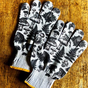 Op de afbeelding: Een paar witte werkhandschoenen met een zwarte grafische print van diverse paddenstoelen, varens en kikkers. De handschoenen hebben een geribbelde gebreide boord met een gele rand bij de opening voor een goede pasvorm tijdens het werk.