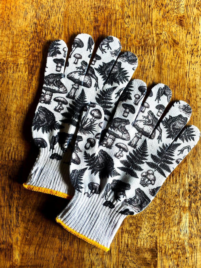 Op de afbeelding: Een paar witte werkhandschoenen met een zwarte grafische print van diverse paddenstoelen, varens en kikkers. De handschoenen hebben een geribbelde gebreide boord met een gele rand bij de opening voor een goede pasvorm tijdens het werk.