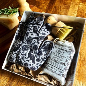 Op de afbeelding: Een geschenkdoos met een paar zwart-wit bedrukte handschoenen met raven- en bloemenmotief, een houten afwasborstel met gele haren en een stuk Nevermore Dirt Oats & Honey zeep met een gewicht van 114g/4 oz.