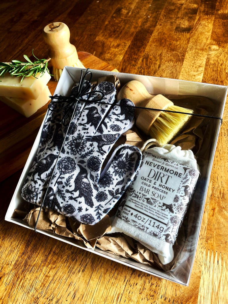 Op de afbeelding: Een geschenkdoos met een paar zwart-wit bedrukte handschoenen met raven- en bloemenmotief, een houten afwasborstel met gele haren en een stuk Nevermore Dirt Oats & Honey zeep met een gewicht van 114g/4 oz.