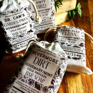 Op de afbeelding: Drie rechthoekige blokken koudgeperste zeep, elk verpakt in een cr&egrave;mekleurig stoffen zakje met trekkoord. De labels bevatten zwarte tekst en botanische illustraties, met de namen Dr. Caw's Garden Paws, Dirty Bird, Toad-Ally Clean en Nevermore Dirt, elk met een gewicht van 114g.