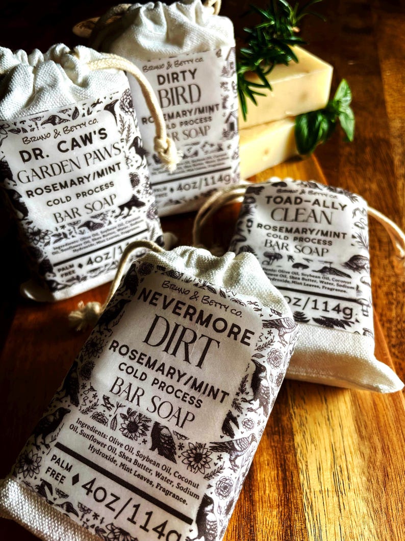 Op de afbeelding: Drie rechthoekige blokken koudgeperste zeep, elk verpakt in een cr&egrave;mekleurig stoffen zakje met trekkoord. De labels bevatten zwarte tekst en botanische illustraties, met de namen Dr. Caw's Garden Paws, Dirty Bird, Toad-Ally Clean en Nevermore Dirt, elk met een gewicht van 114g.