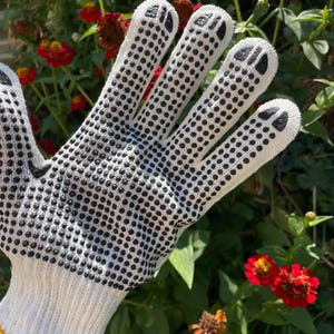 Op de afbeelding: Een witte gebreide handschoen met een zwart stippenpatroon voor grip op de handpalm en vingers, met zwarte pootafdrukken op de toppen van elke vinger. De handschoen heeft een gele geribbelde boord bij de pols voor een goede pasvorm.